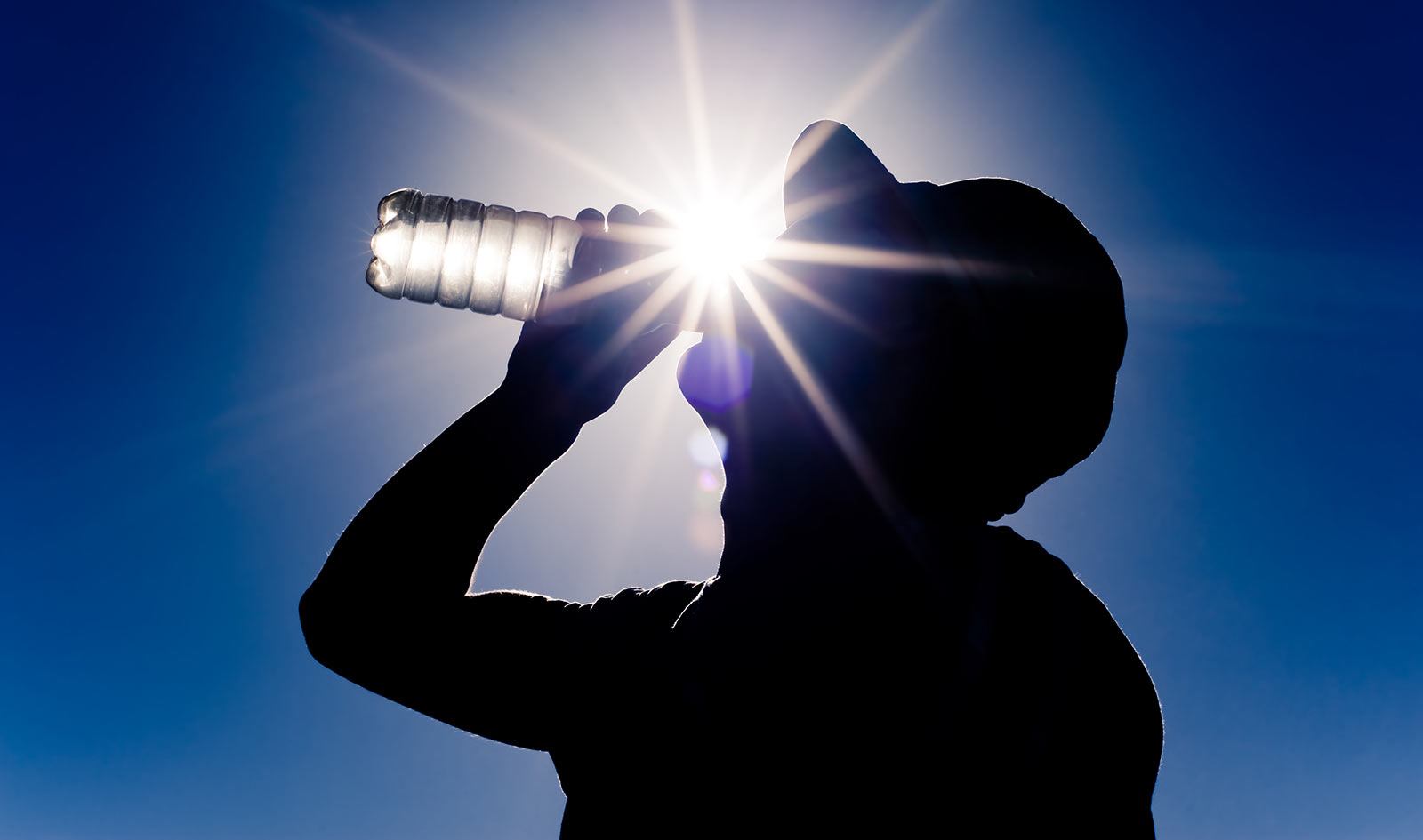 A person wearing a baseball cap drinking water in front of the sun