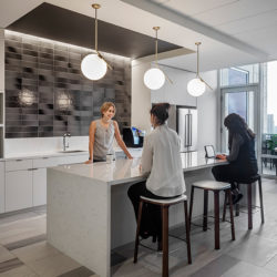 Three people in an office breakroom conversing around a kitchen island with barstools.