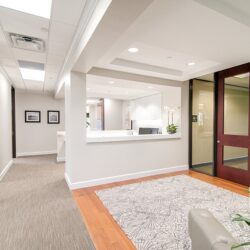 An office with tall ceilings, white walls, a glass door entrance on the right and 3 brown wooden doors on the left